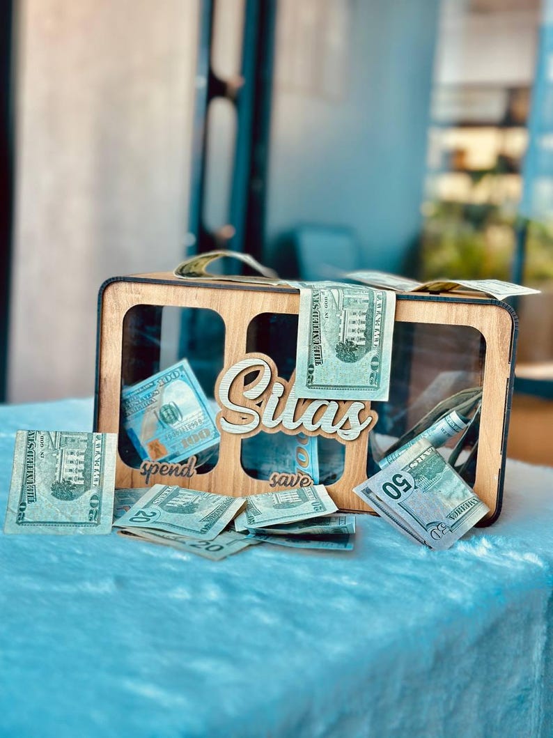 May include: A rectangular wooden money box with three clear acrylic windows and the name Silas in script lettering. The box features labels for spend and save, and is filled with various United States paper currency bills including twenties and fifties.