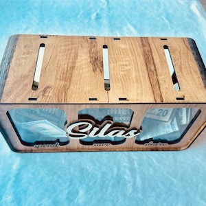 May include: A rectangular wooden money box with three clear acrylic front panels, featuring the name Silas in script lettering. The box has three top slots and labels for spend, save, and share below each section, containing US paper currency.