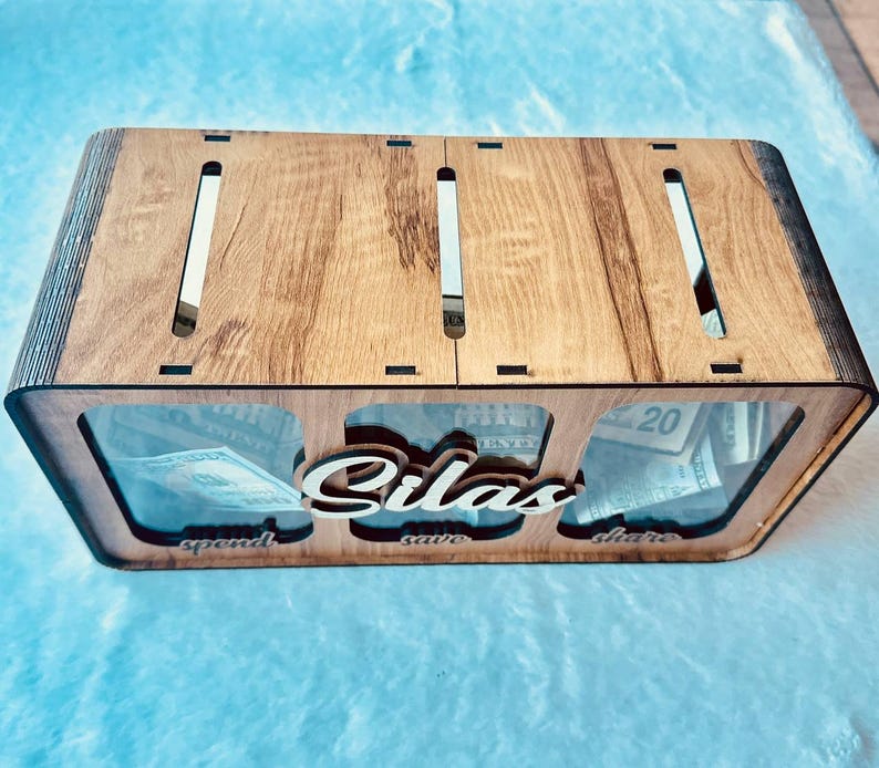 May include: A rectangular wooden money box with three clear acrylic front panels, featuring the name Silas in script lettering. The box has three top slots and labels for spend, save, and share below each section, containing US paper currency.