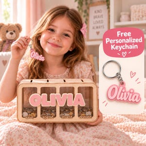 Könnte beinhalten: Eine rechteckige Spardose aus Holz mit vier durchsichtigen Fächern, beschriftet mit save, spend, invest und donate, sowie dem Namen Olivia in rosa Buchstaben. Ein passender rosa Schlüsselanhänger mit dem Namen Olivia ist als Gratisartikel enthalten.