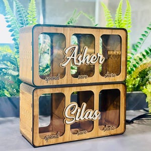 May include: Two stacked rectangular wooden money boxes with clear fronts. Each box features three compartments labelled spend, save, and share. The top box has the name Asher in white script, and the bottom box has the name Silas in white script.