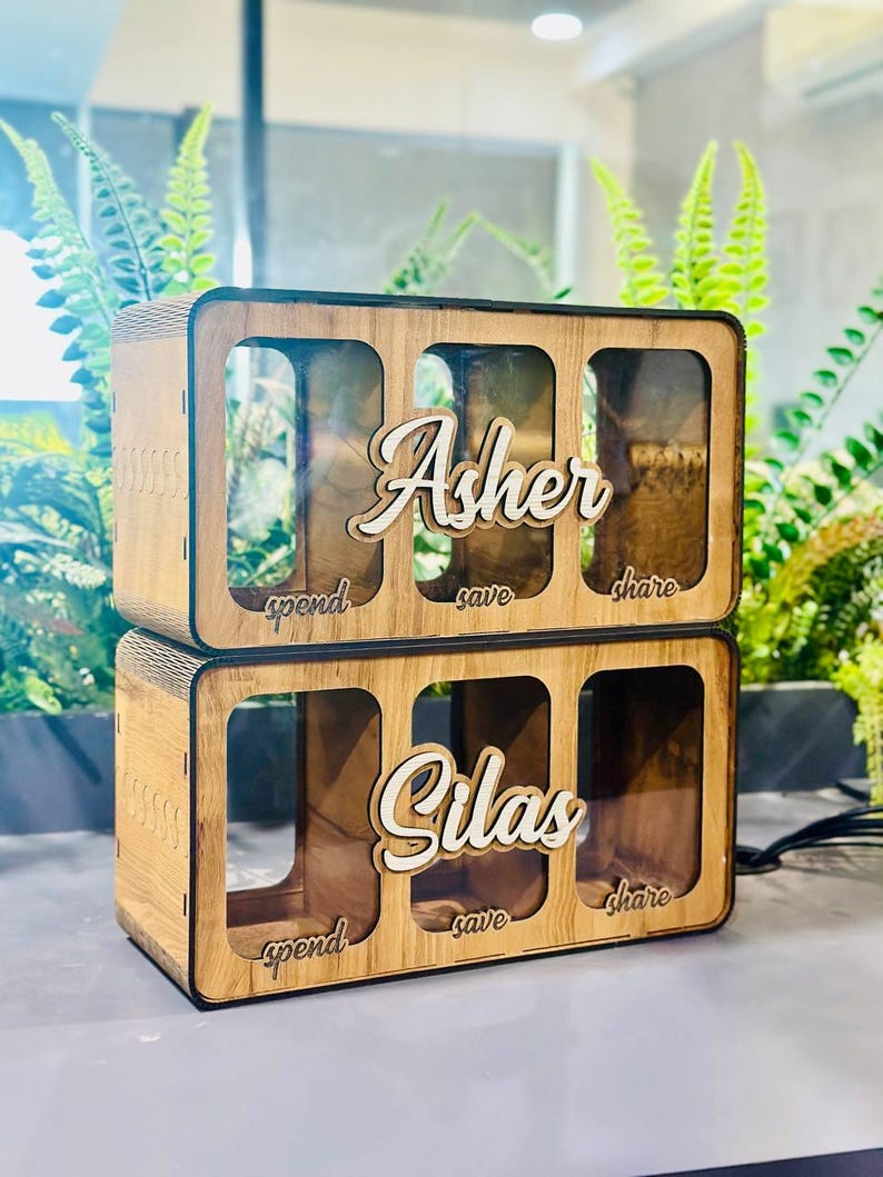 May include: Two stacked rectangular wooden money boxes with clear fronts. Each box features three compartments labelled spend, save, and share. The top box has the name Asher in white script, and the bottom box has the name Silas in white script.
