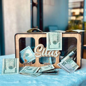 May include: A rectangular wooden money box with three clear acrylic windows, featuring the name Silas in white script lettering. Below the windows, the words spend and save are engraved. The box is filled with various United States dollar bills.
