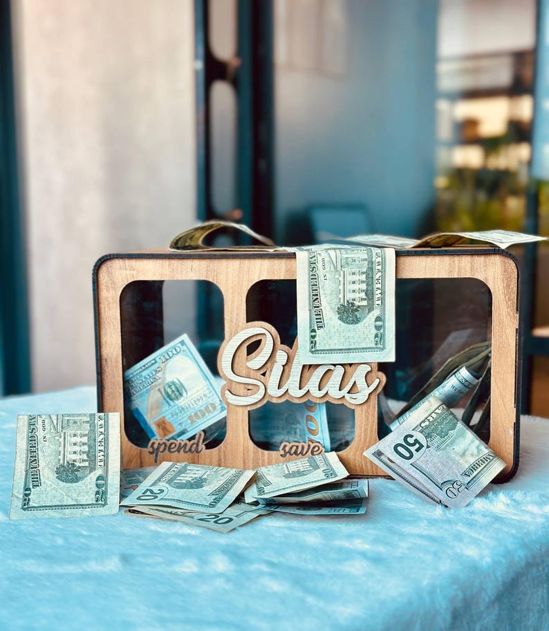 May include: A rectangular wooden money box with three clear acrylic windows, featuring the name Silas in white script lettering. Below the windows, the words spend and save are engraved. The box is filled with various United States dollar bills.