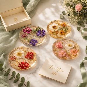 May include: A set of four round resin coasters embedded with pressed flowers and gold flakes, accompanied by an open cream-colored gift box and a small card with the gold-printed text For You, arranged on a white surface with green fabric.