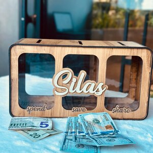 May include: A rectangular wooden money box with three clear acrylic compartments labelled spend, save, and share. The name Silas is cut out in wood and attached to the centre. US dollar bills are placed in front of the box on a white surface.