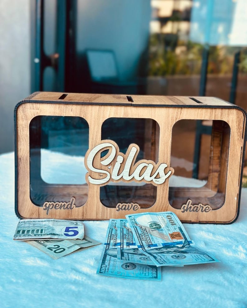 May include: A rectangular wooden money box with three clear acrylic compartments labelled spend, save, and share. The name Silas is cut out in wood and attached to the centre. US dollar bills are placed in front of the box on a white surface.