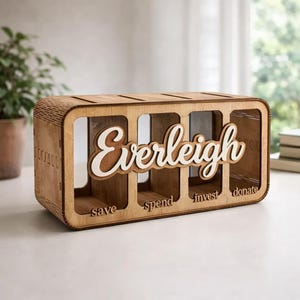 Może przedstawiać: Prostokątna drewniana skarbonka z czterema przezroczystymi przegródkami. Z przodu znajduje się imię Everleigh wypisane białym pismem odręcznym. Pod każdą przegródką wygrawerowano małą czcionką słowa save, spend, invest oraz donate.