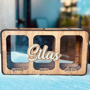 May include: A rectangular wooden money box featuring three clear acrylic compartments. The front displays the name Silas in raised script lettering, with the words spend, save, and share engraved below each corresponding section.