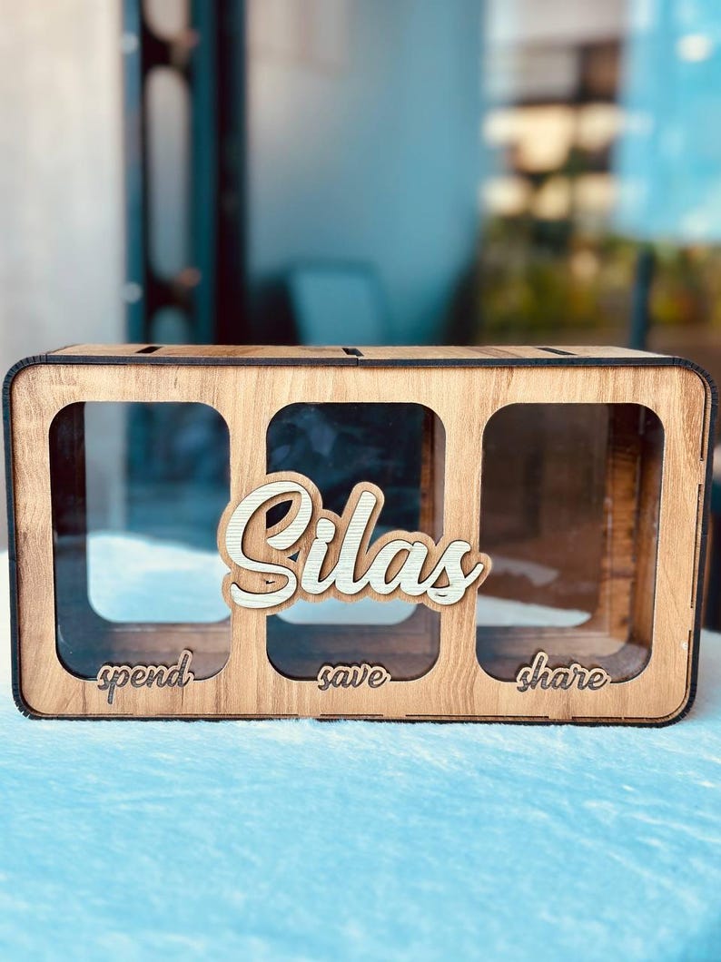 May include: A rectangular wooden money box featuring three clear acrylic compartments. The front displays the name Silas in raised script lettering, with the words spend, save, and share engraved below each corresponding section.