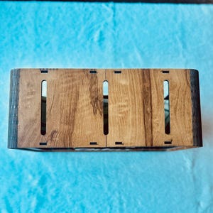May include: A rectangular wooden box with a natural wood grain finish, featuring three vertical rectangular cutouts on the top panel and dark, charred edges from laser cutting, showing a hollow interior through the slots.