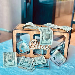 May include: A rectangular wooden money box with three clear acrylic front panels. The name Silas is cut out in wood script on the front. Below the name, the words spend and save are engraved. US dollar bills are placed inside and around the box.