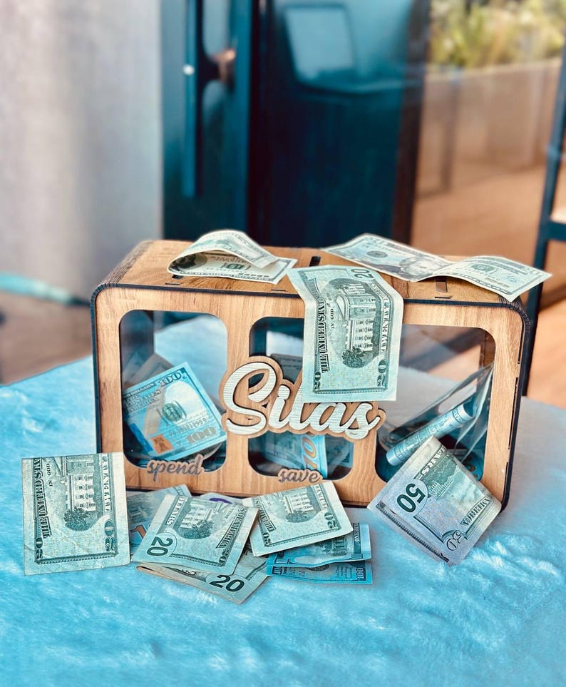 May include: A rectangular wooden money box with three clear acrylic front panels. The name Silas is cut out in wood script on the front. Below the name, the words spend and save are engraved. US dollar bills are placed inside and around the box.