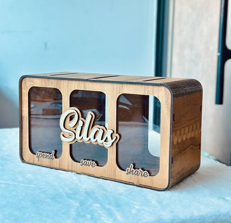 May include: A rectangular wooden money box with three clear acrylic front panels. The name Silas is written in large script letters above the centre panel. Below each panel, the words spend, save, and share are written in small script letters.