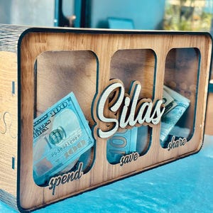 May include: A rectangular wooden money box with three clear acrylic front panels. The name Silas is cut out in wood script lettering in the centre. Below each panel, the words spend, save, and share are engraved. US dollar bills are visible inside.