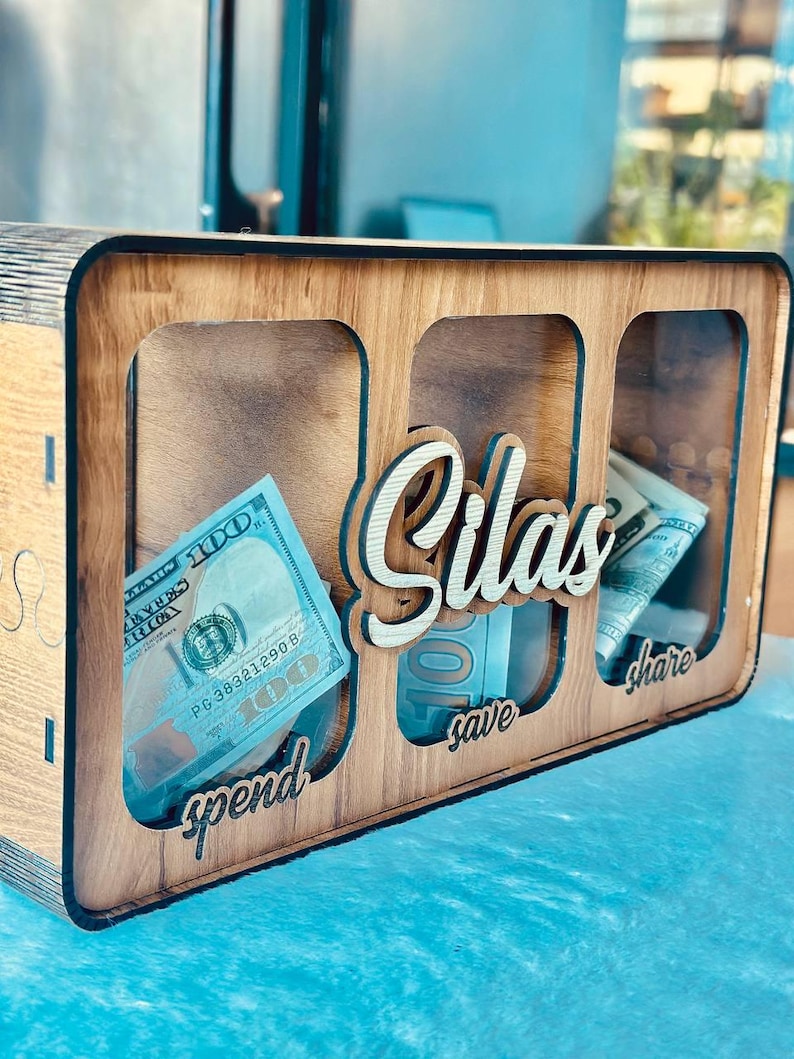 May include: A rectangular wooden money box with three clear acrylic front panels. The name Silas is cut out in wood script lettering in the centre. Below each panel, the words spend, save, and share are engraved. US dollar bills are visible inside.