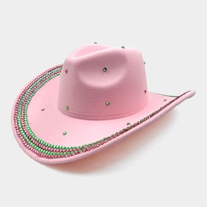 May include: A pink cowboy hat with a curved brim and a rounded crown. The hat is decorated with green and pink rhinestones around the brim and scattered across the crown. A fun and fashionable accessory.