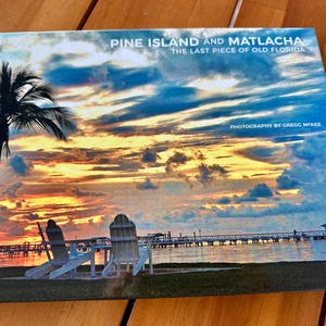 Könnte beinhalten: Ein Buch mit dem Titel "Pine Island and Matlacha: The Last Piece of Old Florida" mit einem Foto eines Sonnenuntergangs über dem Wasser mit zwei Stühlen am Strand. Der Text "Photography by Gregg McKee" befindet sich am unteren Rand des Covers.