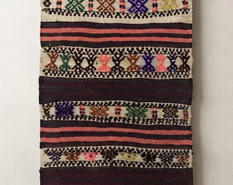 Puede incluir: Un tapiz rectangular tejido con rayas horizontales en color morado oscuro y rosa apagado. El tejido incluye bandas de tela color crema decoradas con patrones geométricos repetitivos en verde, azul, amarillo y rosa, rematado con flecos.