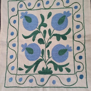 May include: A white fabric square with a blue and green embroidered floral design. The design features four stylized flowers with green leaves and stems. The flowers are surrounded by a green and blue border.