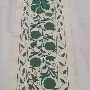 May include: Cream-colored rectangular textile with intricate green embroidery. The design features floral and fruit motifs, including stylized flowers and round shapes, with a border of connected leaves. The embroidery is detailed and creates a textured appearance.