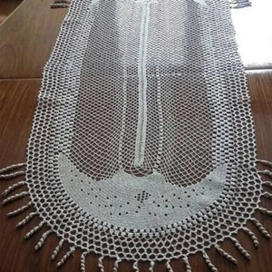 Puede incluir: Un camino de mesa blanco de ganchillo con un borde festoneado y flecos. El camino de mesa tiene un diseño delicado y de encaje.