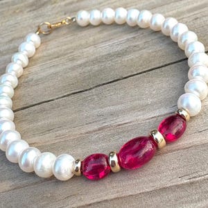 Può includere: Un delicato braccialetto con perle bianche e vivaci pietre preziose rosa. Il braccialetto è impreziosito da metallo color oro e una chiusura sicura. Le perle e le pietre preziose sono infilate insieme, creando un bellissimo gioiello.