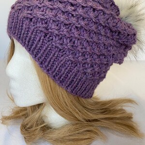 May include: A knitted purple beanie with a textured pattern and a faux fur pom-pom. The hat is made of a soft, warm yarn, perfect for cold weather. The beanie is shown on a mannequin head with blonde hair.