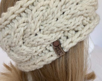 Può includere: Un cerchietto di lana bianco con un motivo a treccia. Il cerchietto ha un'etichetta in pelle marrone con il testo "West Byron" su di essa.