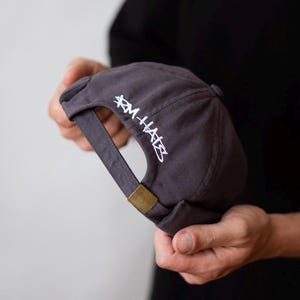 May include: A charcoal gray baseball cap featuring a rolled brim and an adjustable strap with a brass-toned metal buckle. The back of the cap is embroidered with white text that reads RM HATS in a stylized, graffiti-inspired script font.