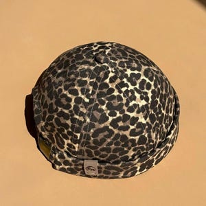 Könnte beinhalten: Ein braun-schwarzer Beanie-Hut mit Leopardenmuster. Der Hut ist aus weichem Stoff gefertigt und hat eine kurze Krempe. An der Seite des Hutes ist ein kleines Etikett angebracht.