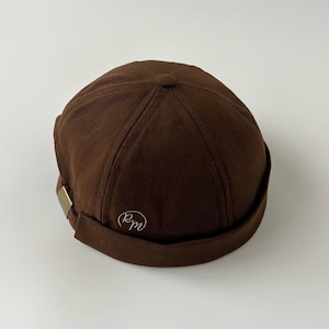 May include: A brown skull cap with a rolled brim and a small, embroidered design on the side. The cap has a simple, classic design with a metal clasp detail. The cap is made of a sturdy fabric.