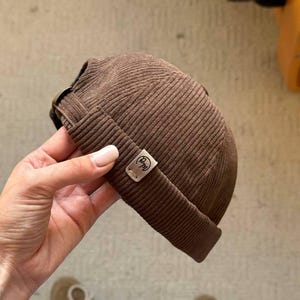 May include: A brown corduroy beanie hat with a small rectangular tag. The hat features a rolled-up brim and a metal buckle. The tag displays the letters "Pm". The hat is a neutral colour and suitable for anyone.
