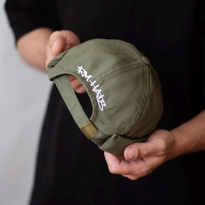 May include: An olive green brimless cap featuring a rolled edge and an adjustable strap with a brass-toned metal buckle. The back of the cap is embroidered with white text that reads RM HATS in a stylized, angular font.