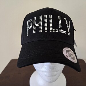 May include: Black baseball cap with the word "PHILLY" spelled out in white rhinestones. The cap has a curved brim and a tag attached. The cap is displayed on a white mannequin head.
