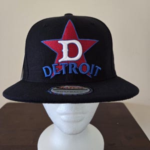 Peut inclure: Casquette noire avec une étoile rouge et un logo "D" blanc. Le mot "DETROIT" est brodé en bleu en dessous. La casquette a une visière plate et une fermeture à pression.