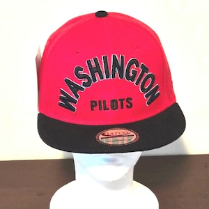 Peut inclure: Une casquette de baseball rouge et noire avec le texte "WASHINGTON PILOTS" en lettres noires sur le devant. La casquette a une visière noire et une étiquette noire "HATCO SNAPBACK" au dos.