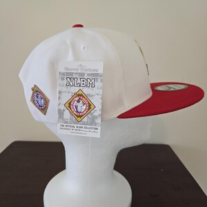Puede incluir: Gorra de b&eacute;isbol blanca con visera roja y un peque&ntilde;o parche en el lateral. La gorra tiene una etiqueta adjunta que dice "Discover Greatness NLBM The Official NLBM Collection Exclusively by New Era".