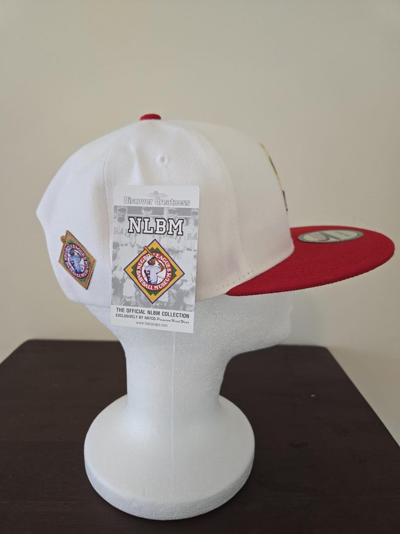 Puede incluir: Gorra de b&eacute;isbol blanca con visera roja y un peque&ntilde;o parche en el lateral. La gorra tiene una etiqueta adjunta que dice "Discover Greatness NLBM The Official NLBM Collection Exclusively by New Era".