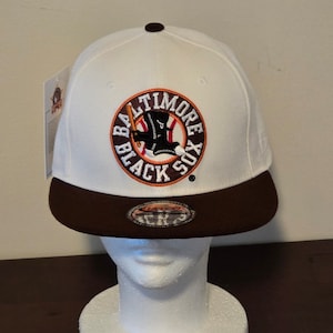 Peut inclure: Casquette de baseball blanche avec une visière marron. L'avant de la casquette présente un logo circulaire avec le texte "Baltimore Black Sox" en lettres orange et noires. Le logo comprend également une image de dessin animé d'un joueur de baseball portant un uniforme noir et tenant une batte.