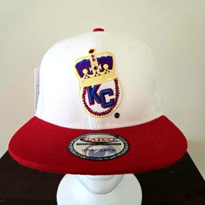 Puede incluir: Gorra de b&eacute;isbol blanca y roja con una corona dorada y "KC" bordado en la parte delantera. La gorra tiene una etiqueta "Hatco Premium Headwear Snapback" en la parte inferior de la visera.