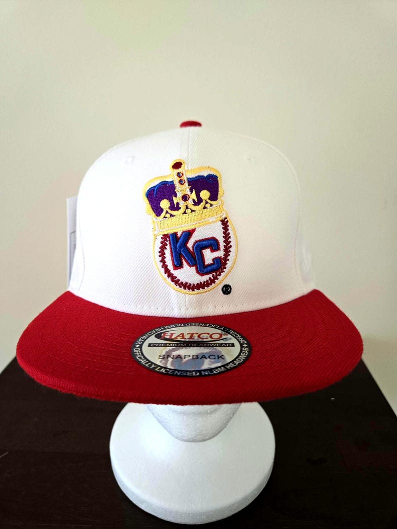 Puede incluir: Gorra de b&eacute;isbol blanca y roja con una corona dorada y "KC" bordado en la parte delantera. La gorra tiene una etiqueta "Hatco Premium Headwear Snapback" en la parte inferior de la visera.