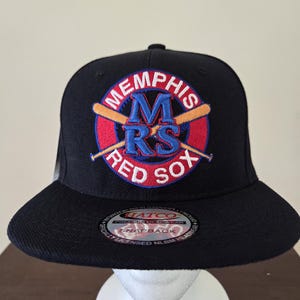 Peut inclure: Casquette de baseball noire avec un logo brodé rouge, blanc et bleu qui dit "Memphis Red Sox" avec un grand "M" et des bâtons de baseball croisés.