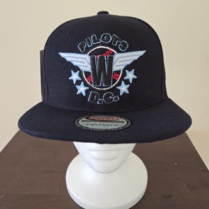 Peut inclure: Casquette de baseball bleu marine avec l'inscription "PILOTS D.C." brodée en blanc, rouge et bleu clair. La casquette présente un logo "W" avec des ailes et des étoiles. Elle a une visière plate et une fermeture à pression.