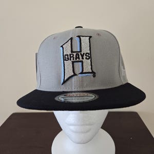Peut inclure: Une casquette de baseball grise et noire avec un "H" brodé en bleu et blanc et le mot "GRAYS" sur le devant.