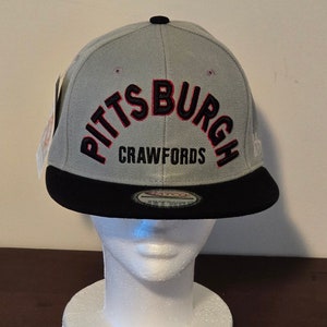 Peut inclure: Casquette de baseball grise et noire avec un contour rouge autour du mot "Pittsburgh" et les mots "Crawfords" en noir en dessous. La casquette est sur une tête de mannequin blanche.