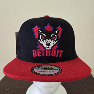 Peut inclure: Casquette snapback noire et rouge à visière plate. Le chapeau présente un logo Detroit avec un design de loup en blanc, rouge et bleu. Le mot "DETROIT" est brodé en rouge. Le chapeau a un autocollant "HATCO".