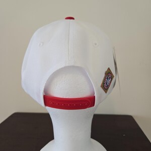 Puede incluir: Gorra de b&eacute;isbol blanca con visera roja y un peque&ntilde;o parche en la parte posterior. El parche tiene un logotipo con un dise&ntilde;o rojo, blanco y azul.