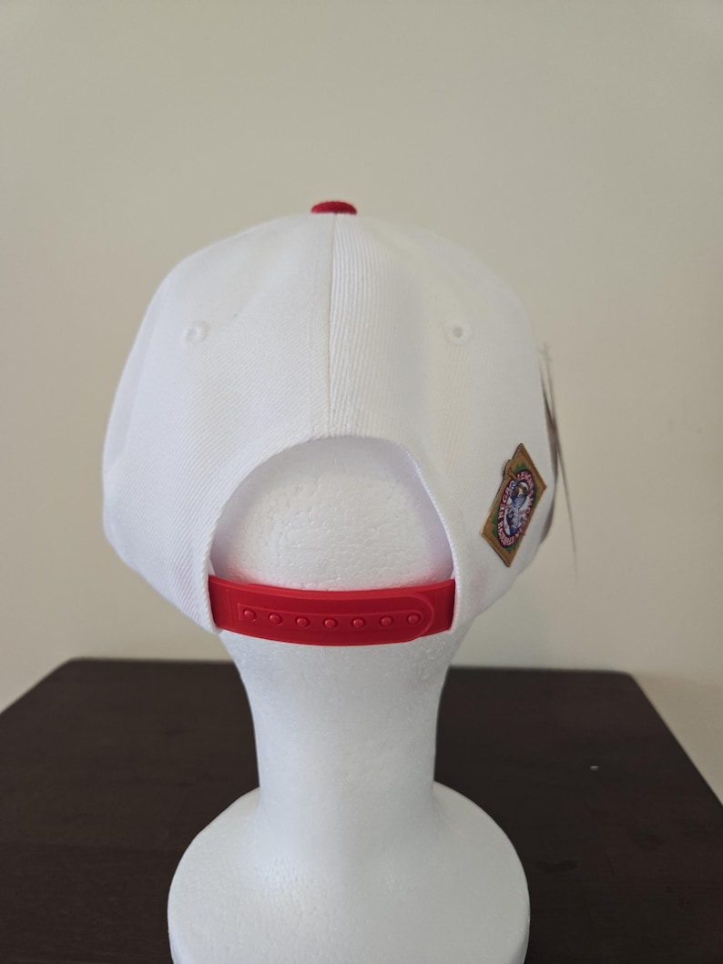 Puede incluir: Gorra de b&eacute;isbol blanca con visera roja y un peque&ntilde;o parche en la parte posterior. El parche tiene un logotipo con un dise&ntilde;o rojo, blanco y azul.