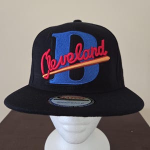 Peut inclure: Casquette de baseball noire avec le mot "Cleveland" en script rouge sur un "B" bleu et une batte de baseball. La casquette a une visière plate et une fermeture à pression.